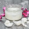 A jar of coconut candle wax with a tea spoon full of coconut scooped from the jar. A few purple flower petals surrounds the jar