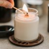 A hand hold a match over a wick, lighting a candle, ready to set its wax memory pool for its first burn. It sits on a brown coaster