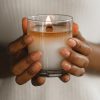 A woman holds a candle with two hands wrapped around the glass. The wax has a full melt pool with a wooden wick being used