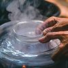 A pan full of boiling water with hands putting a candle jar in the boiling water to melt the candle wax, the boiling water method of removing wax from a candle