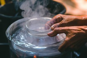 A pan full of boiling water with hands putting a candle jar in the boiling water to melt the candle wax, the boiling water method of removing wax from a candle