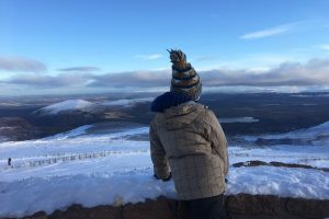 Boy stands at the viewpoint up Cairngorm mountain in the snow-covered hills, clear blue sky in the Highlands of Scotland