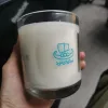 A close-up image of a hand holding a glass candle from Minch Candle Company, showcasing a distinctive Viking ship logo. The candle has large wet spots, where the white wax has visibly pulled away from the glass container, creating uneven adhesion and clear patches. This common occurrence in candle making is exemplified in a real-world product, emphasising the imperfections that can arise despite quality craftsmanship. The background is softly blurred, focusing attention on the candle's details and the aesthetic impact of the wet spots. This image serves as a key visual in a blog post about handling and understanding wet spots in candles.