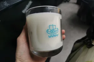 A close-up image of a hand holding a glass candle from Minch Candle Company, showcasing a distinctive Viking ship logo. The candle has large wet spots, where the white wax has visibly pulled away from the glass container, creating uneven adhesion and clear patches. This common occurrence in candle making is exemplified in a real-world product, emphasising the imperfections that can arise despite quality craftsmanship. The background is softly blurred, focusing attention on the candle's details and the aesthetic impact of the wet spots. This image serves as a key visual in a blog post about handling and understanding wet spots in candles.