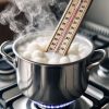 A close-up image showing what happens if you overheat soy wax. The white soy wax is boiling in a pot with steam rising, and a thermometer clearly indicates an overheating temperature. The background is a kitchen setting with a stove, illustrating the candle-making process.