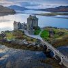 A stunning aerial view of Eilean Donan Castle, located on a small tidal island in the Scottish Highlands, surrounded by calm lochs and connected to the mainland by a stone bridge. The dramatic backdrop features rugged hills and a tranquil sky reflected in the water.