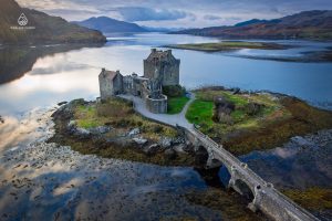 A stunning aerial view of Eilean Donan Castle, located on a small tidal island in the Scottish Highlands, surrounded by calm lochs and connected to the mainland by a stone bridge. The dramatic backdrop features rugged hills and a tranquil sky reflected in the water.