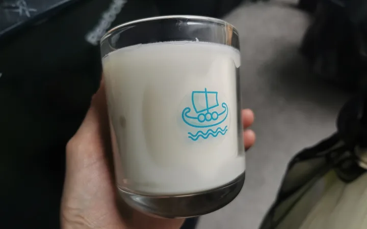 A close-up image of a hand holding a glass candle from Minch Candle Company, showcasing a distinctive Viking ship logo. The candle has large wet spots, where the white wax has visibly pulled away from the glass container, creating uneven adhesion and clear patches. This common occurrence in candle making is exemplified in a real-world product, emphasising the imperfections that can arise despite quality craftsmanship. The background is softly blurred, focusing attention on the candle's details and the aesthetic impact of the wet spots. This image serves as a key visual in a blog post about handling and understanding wet spots in candles.
