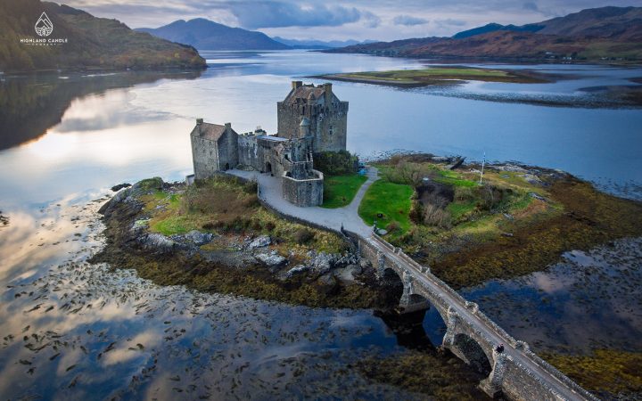 A stunning aerial view of Eilean Donan Castle, located on a small tidal island in the Scottish Highlands, surrounded by calm lochs and connected to the mainland by a stone bridge. The dramatic backdrop features rugged hills and a tranquil sky reflected in the water.