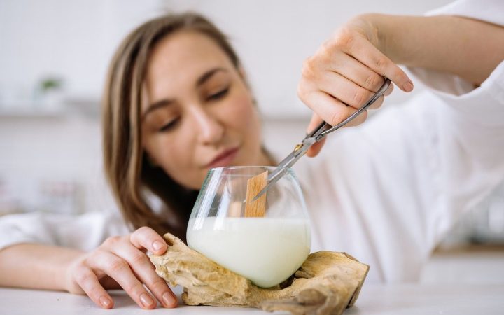 A woman sticks a pair of scissors inside a glass candle jar showing how to trim a candle wick. The wick is a wooden wick in soy wax