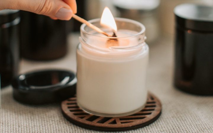 A hand hold a match over a wick, lighting a candle, ready to set its wax memory pool for its first burn. It sits on a brown coaster