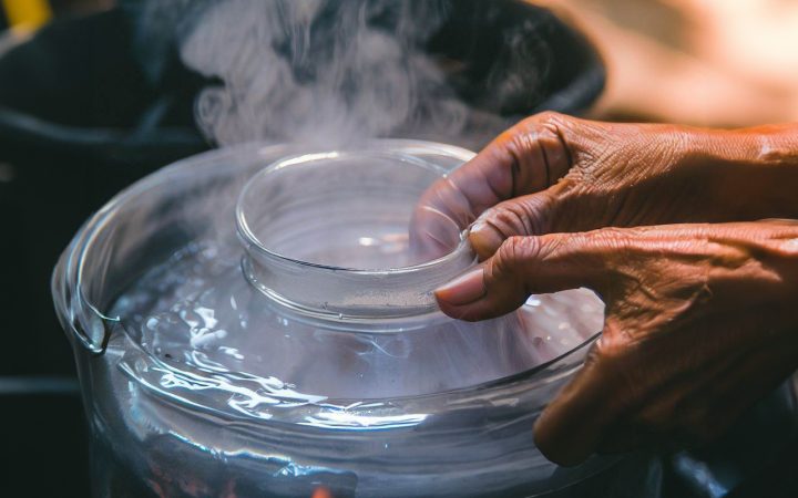 A pan full of boiling water with hands putting a candle jar in the boiling water to melt the candle wax, the boiling water method of removing wax from a candle