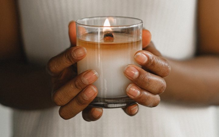A woman holds a candle with two hands wrapped around the glass. The wax has a full melt pool with a wooden wick being used
