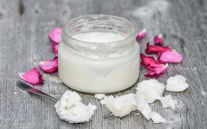 A jar of coconut candle wax with a tea spoon full of coconut scooped from the jar. A few purple flower petals surrounds the jar
